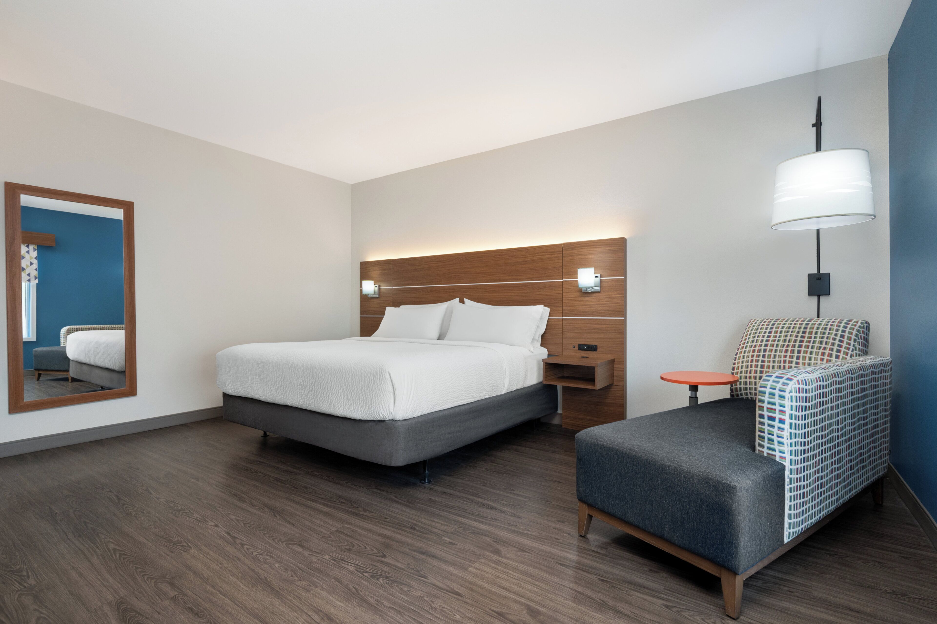 A modern hotel room with a bed, bedside table, chair, and a large mirror.