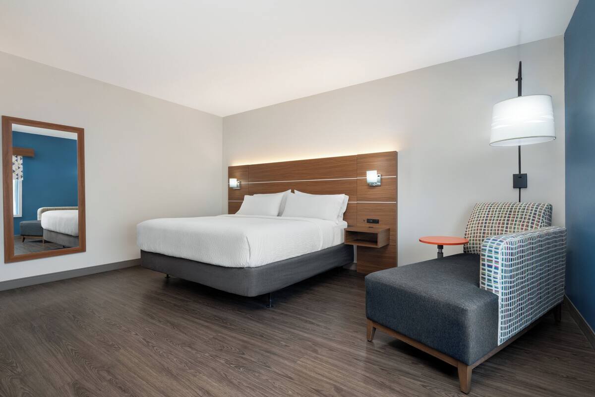 A modern hotel room with a bed, bedside table, chair, and a large mirror.