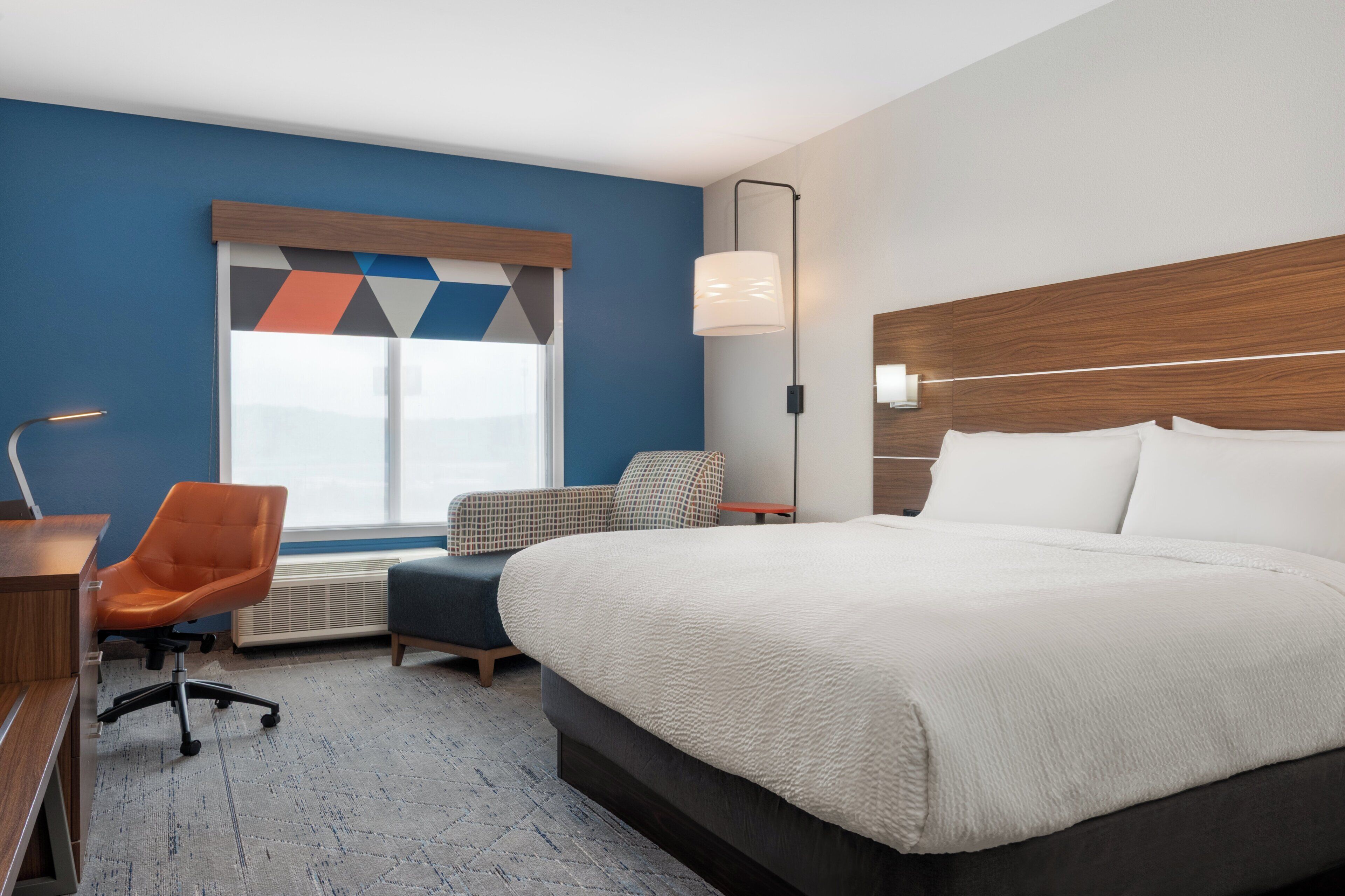 A hotel room with a large bed, a desk, an orange chair, a small couch, a window with geometric curtains, and a wall-mounted lamp.