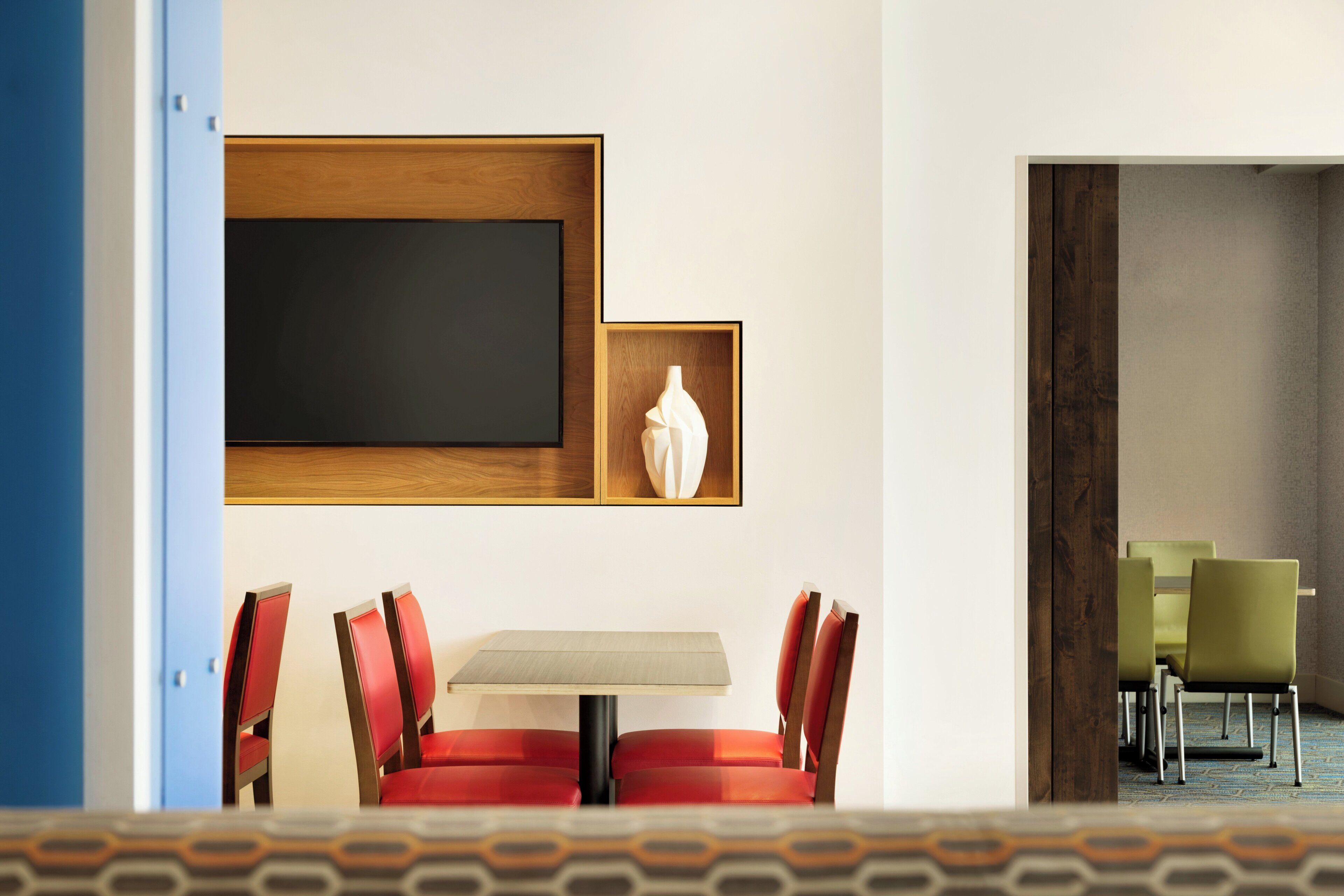 A hotel room with a dining area featuring a table and chairs, a large wooden-framed artwork on the wall, and a view of the adjacent room.