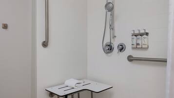 Un baño con baldosas blancas, una ducha con cabezal de ducha manual, un asidero, un perchero con botellas y una mesita con una toalla.