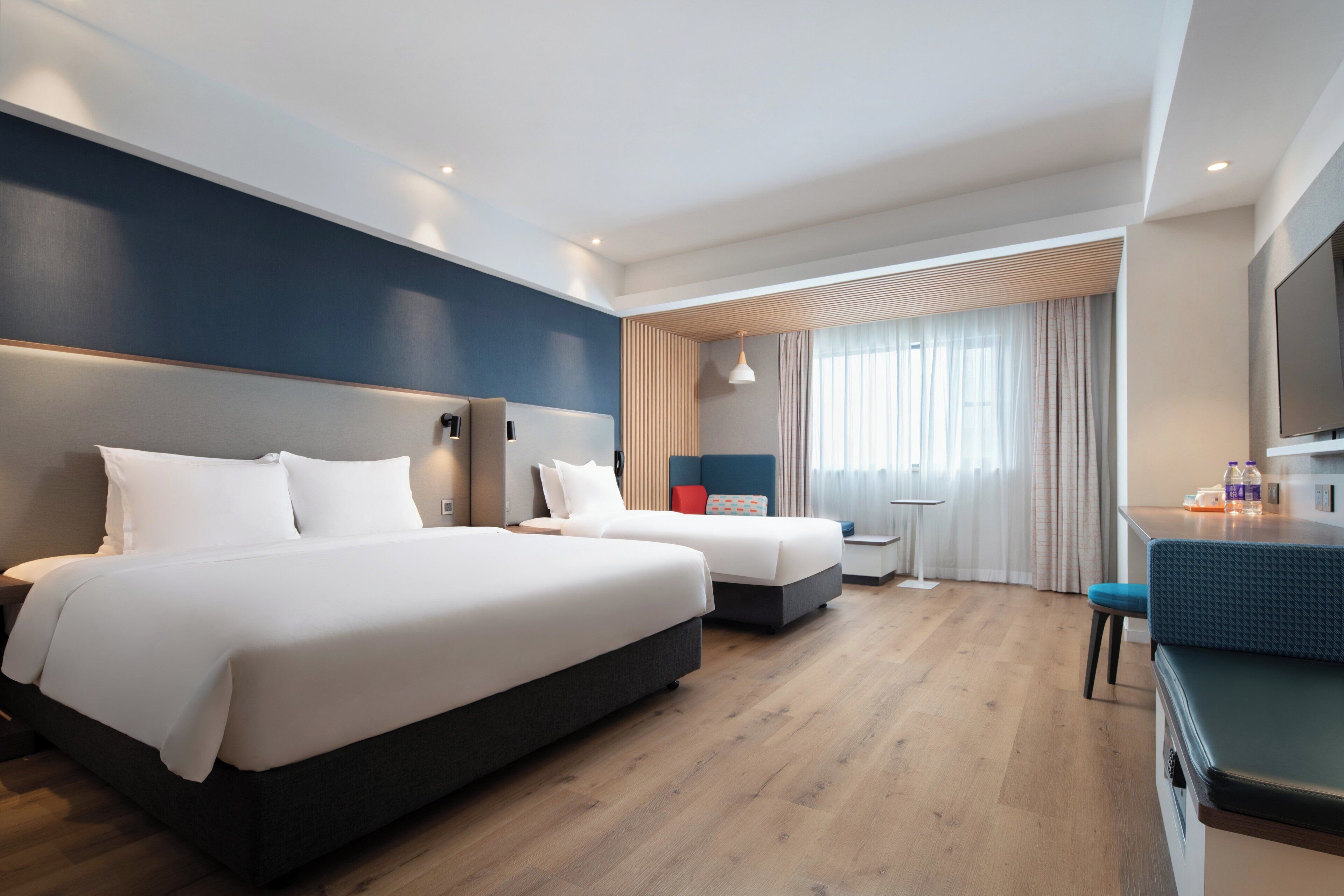 A hotel room with two beds, a desk, a chair, a TV, and a window with curtains.