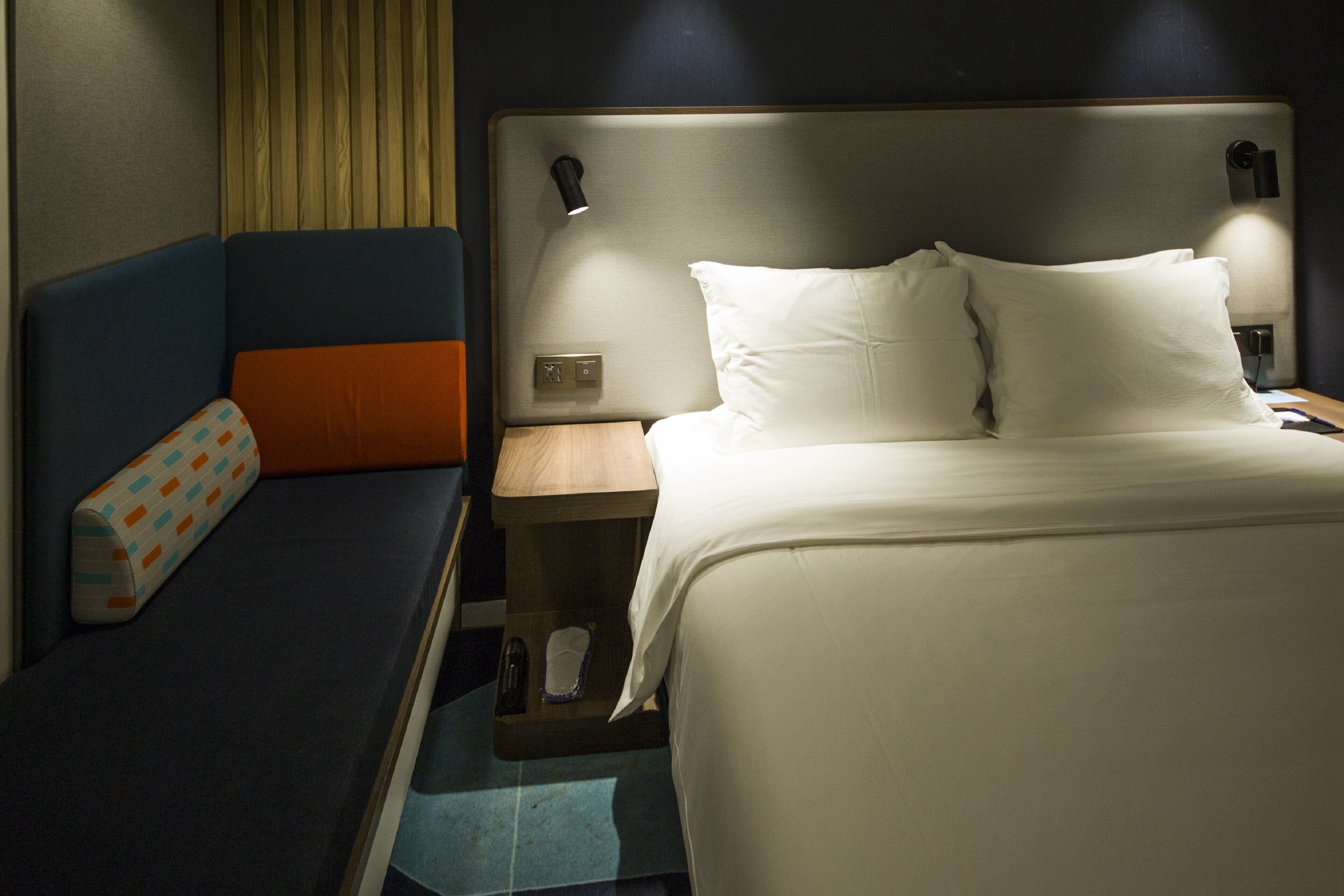 A hotel room with a bed, a bedside table, a nightstand, and a seating area with a bench and pillows.