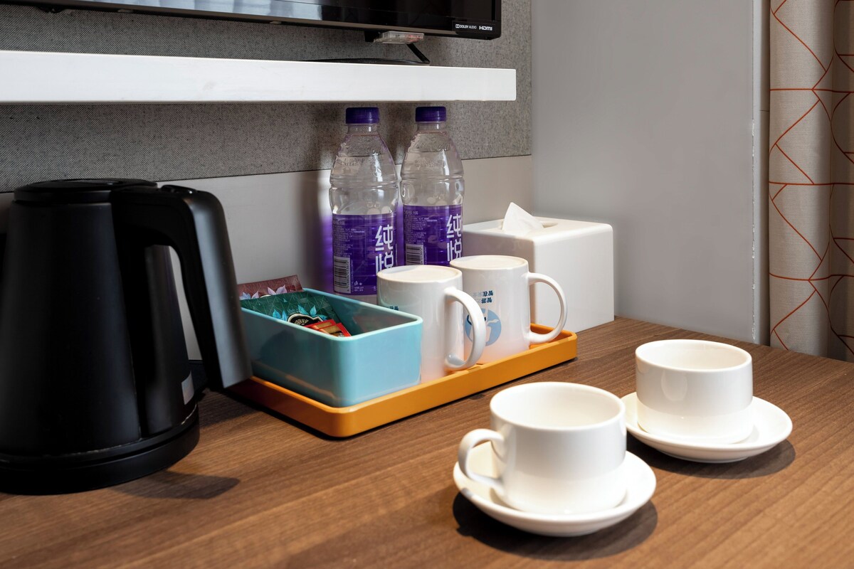 A hotel room with a wooden table, a black kettle, two water bottles, a blue storage box, and white cups and saucers.