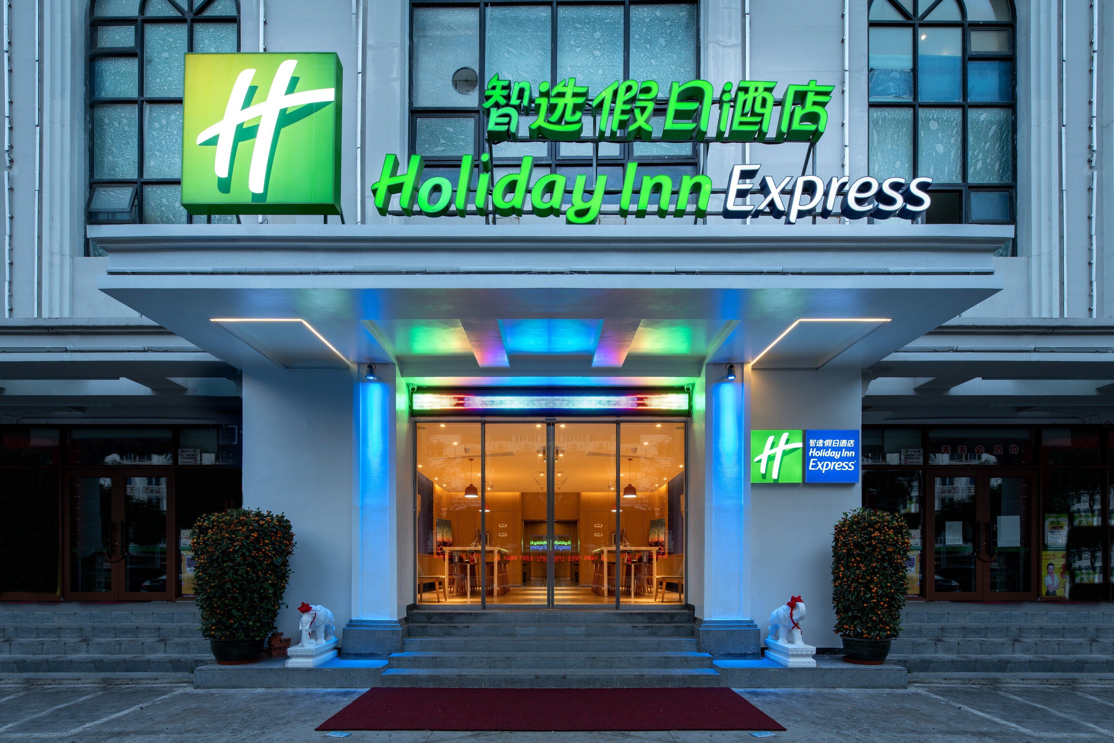 The exterior of a Holiday Inn Express hotel with a prominent green logo, a red carpet, and two potted plants.