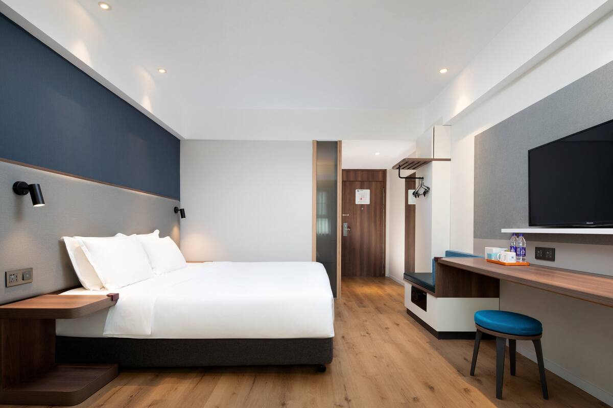 A modern hotel room with a bed, bedside table, a desk with a chair, a TV, and a bathroom door.