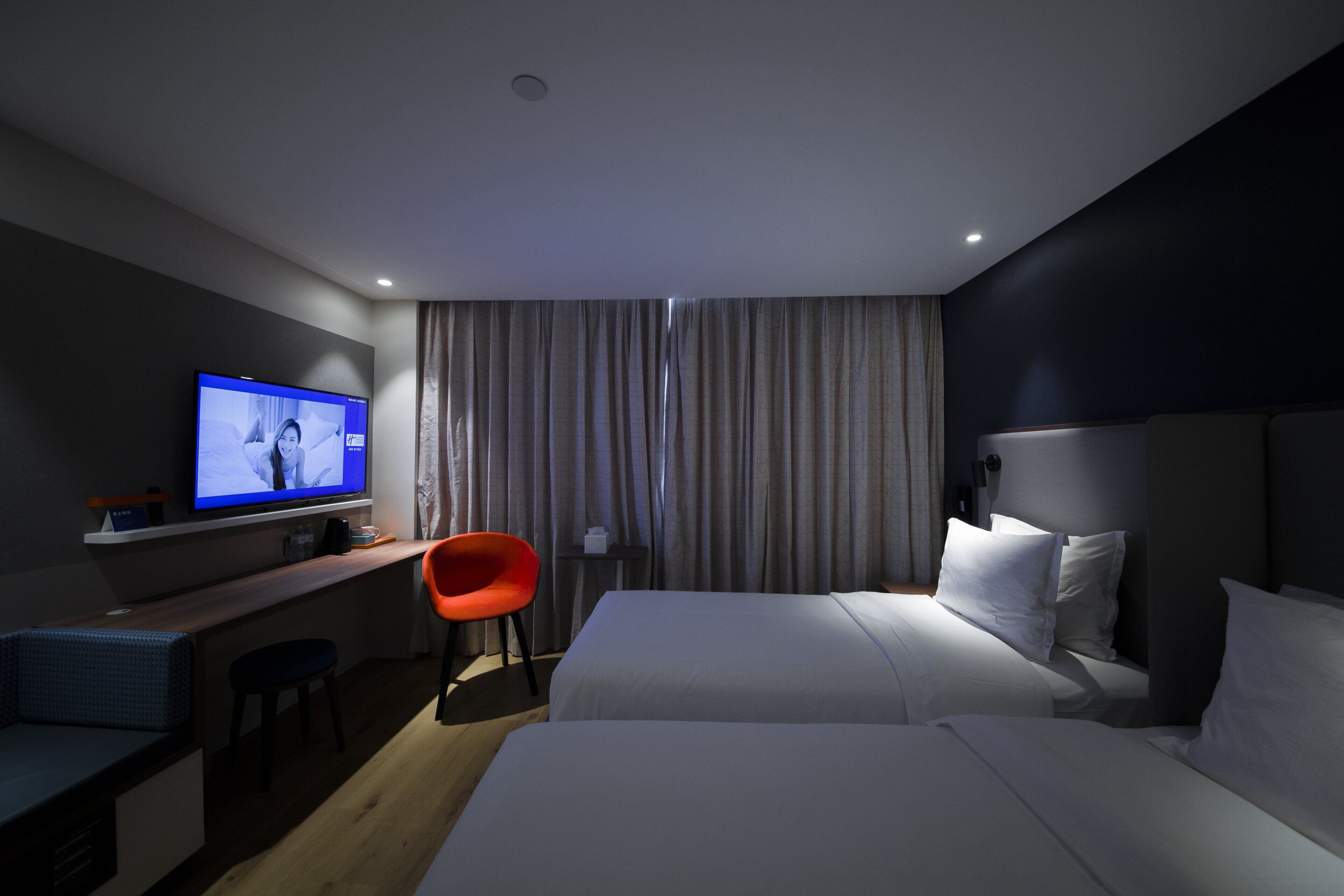 A hotel room with a bed, a desk with a television, a red chair, and a curtain.