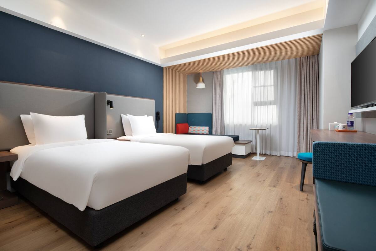 A hotel room with two beds, a desk, a chair, a TV, and a small table.