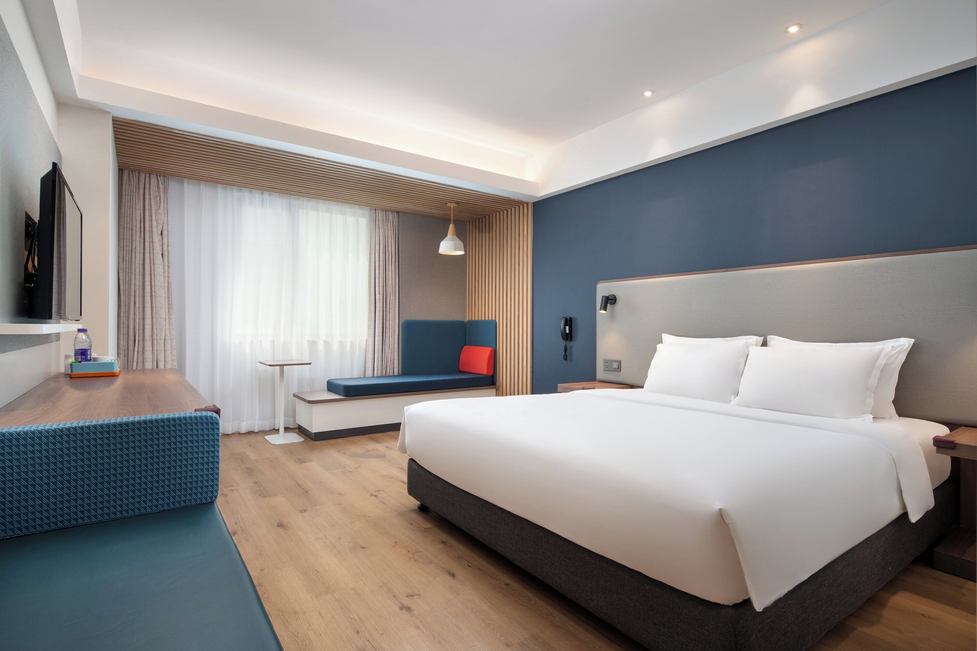 A modern hotel room with a large bed, a seating area, a desk, and a television.