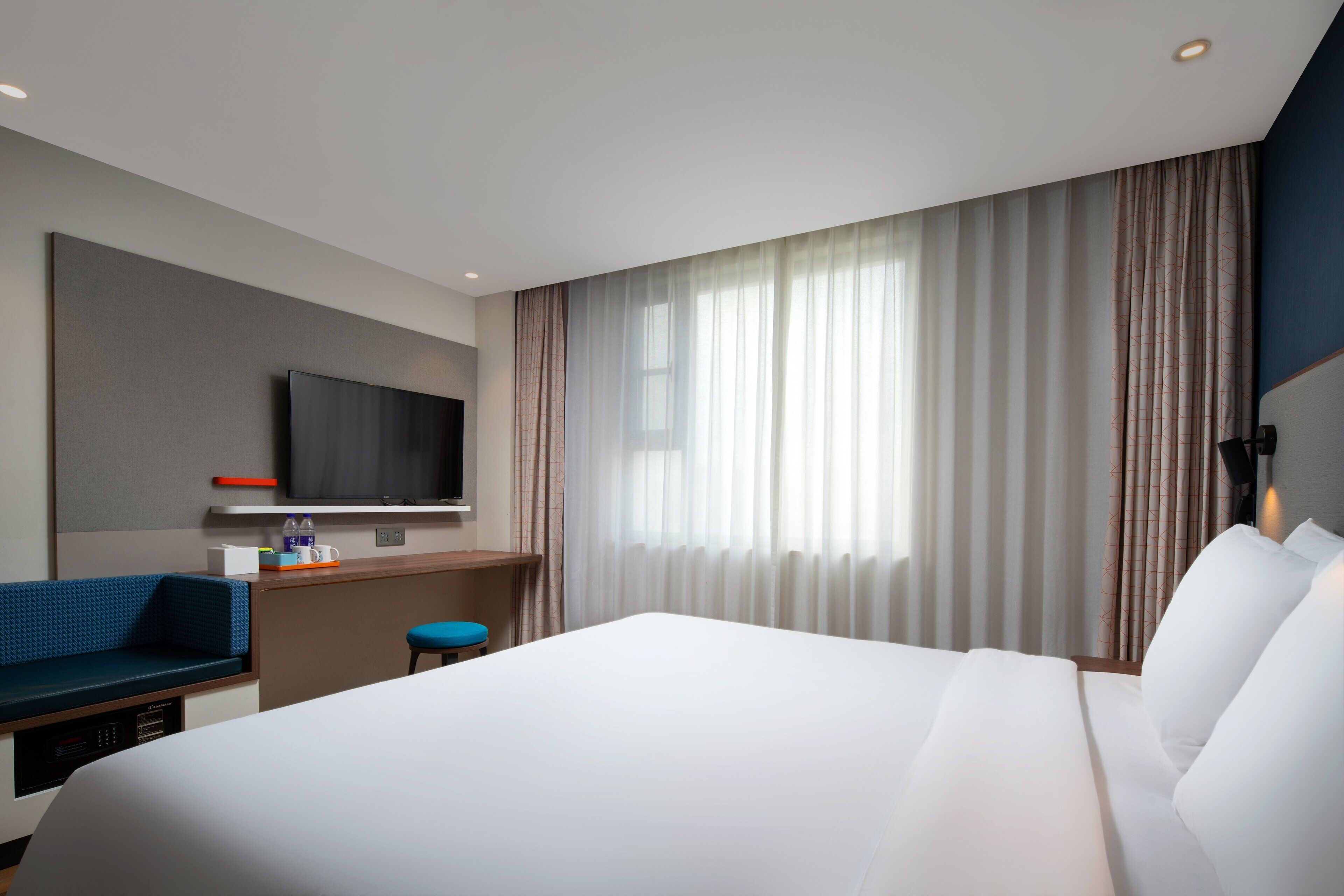Habitación de hotel con una cama grande, un televisor montado en la pared, un escritorio con una silla y una ventana con cortinas.