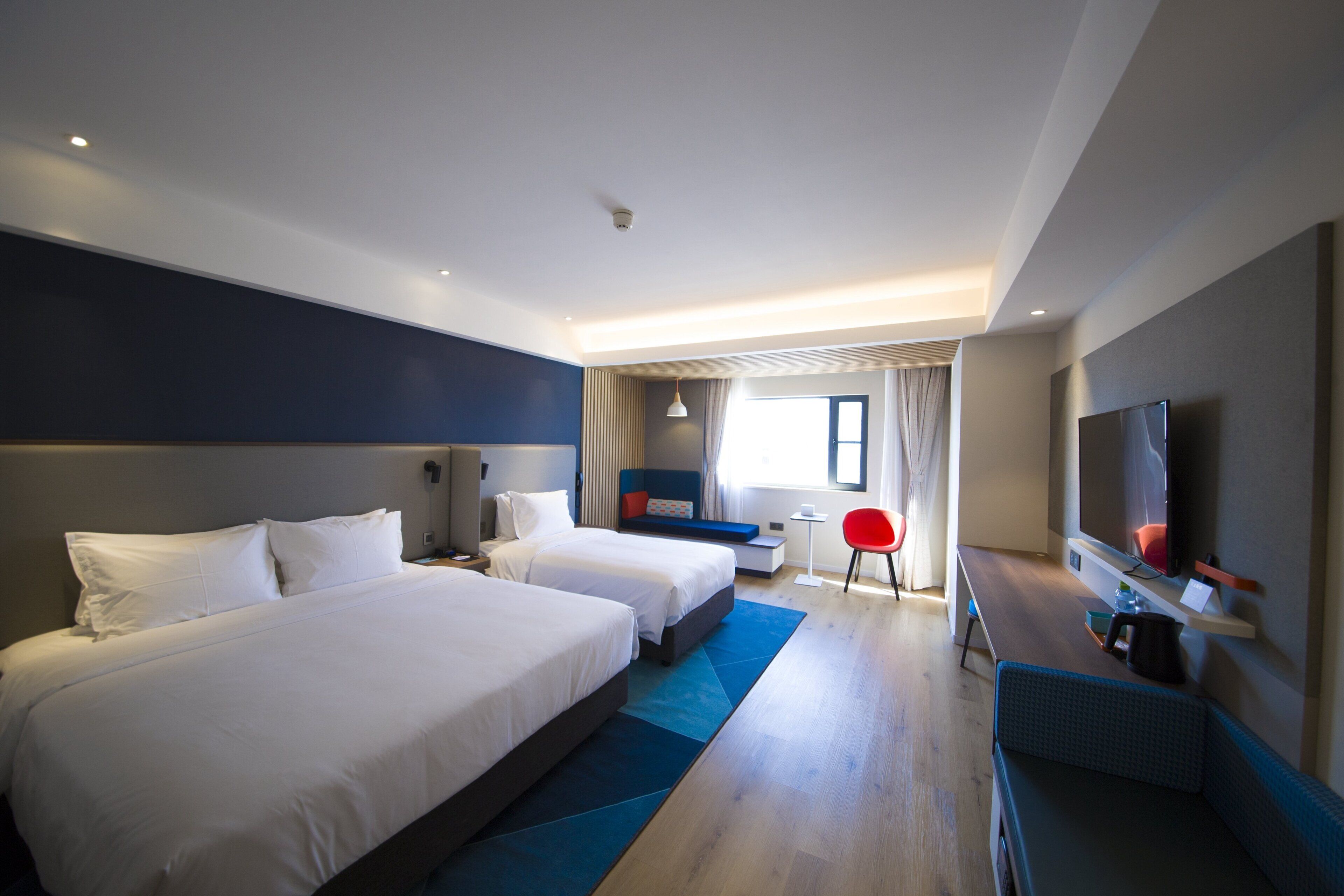 Ein Hotelzimmer mit zwei Betten, einem Schreibtisch, einem Stuhl, einem Fernseher und einem Fenster.