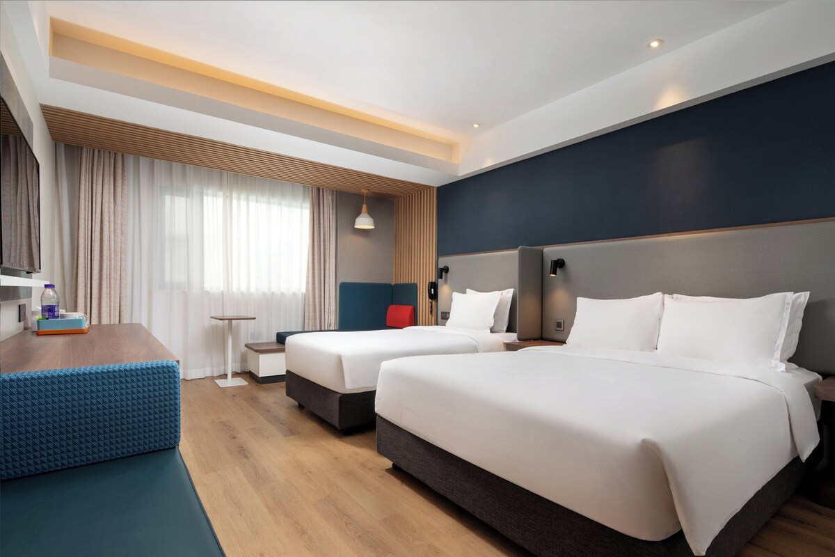 A hotel room with two beds, a wooden floor, a blue bench, and a small table with a bottle and a box.
