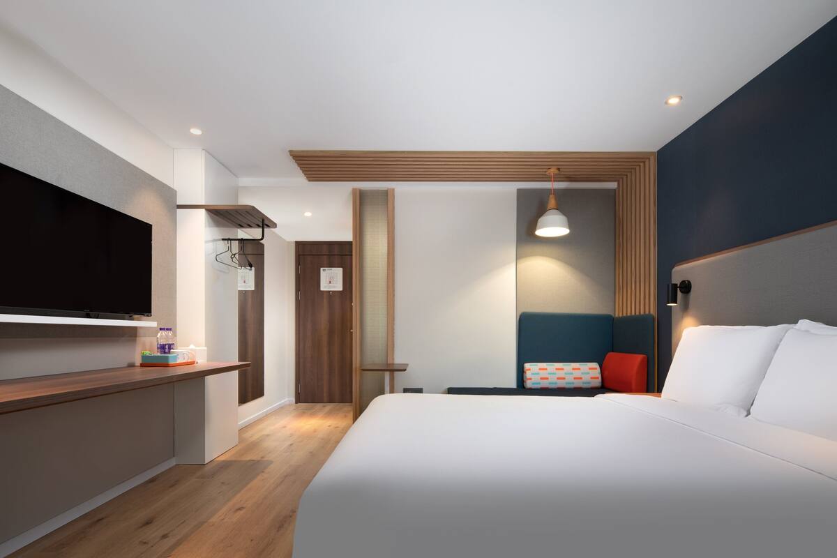 A modern hotel room with a large bed, a flat-screen TV, a desk, and a seating area with a small table and chairs.