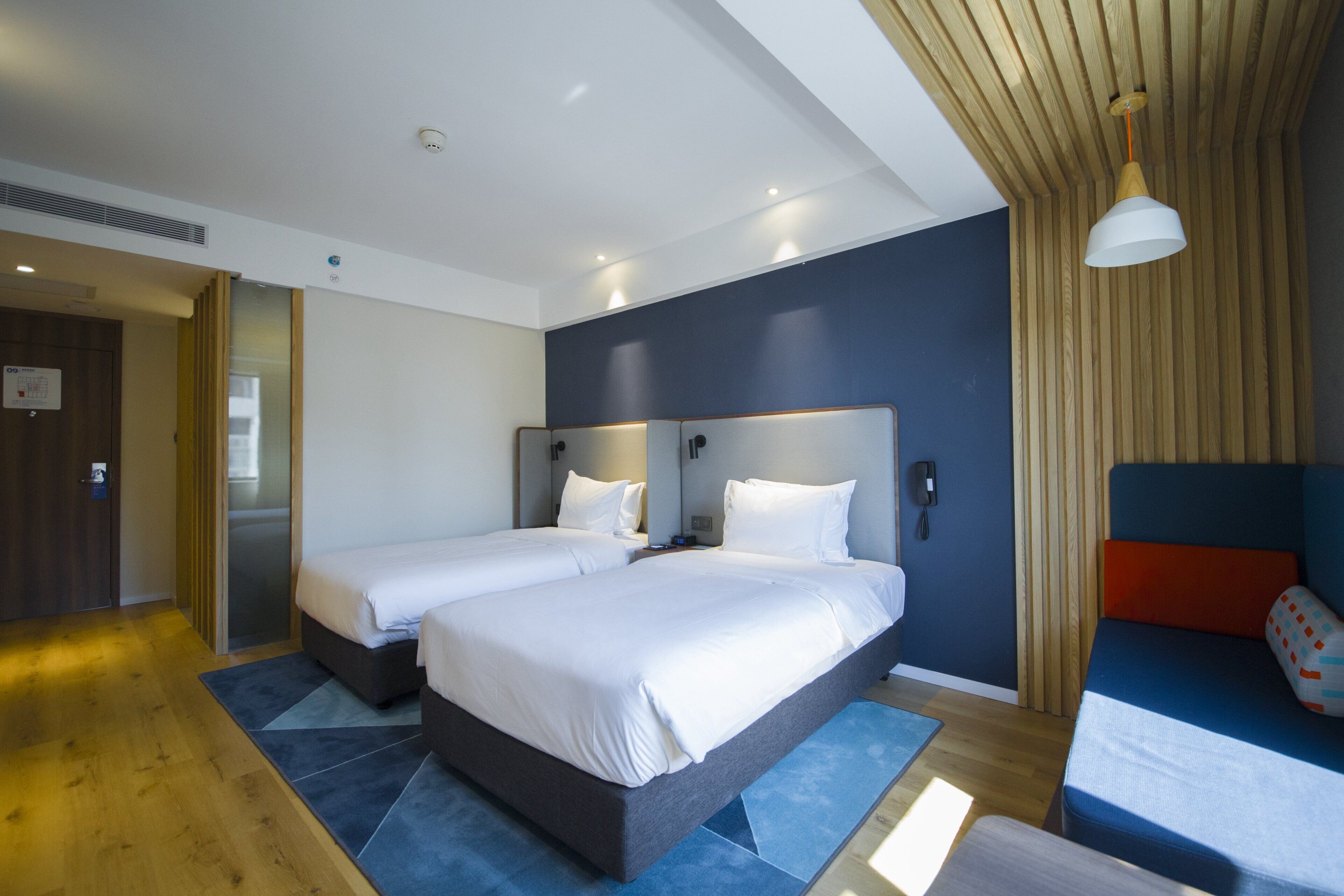 Quarto de hotel moderno com duas camas, teto de madeira e uma parede azul decorativa.