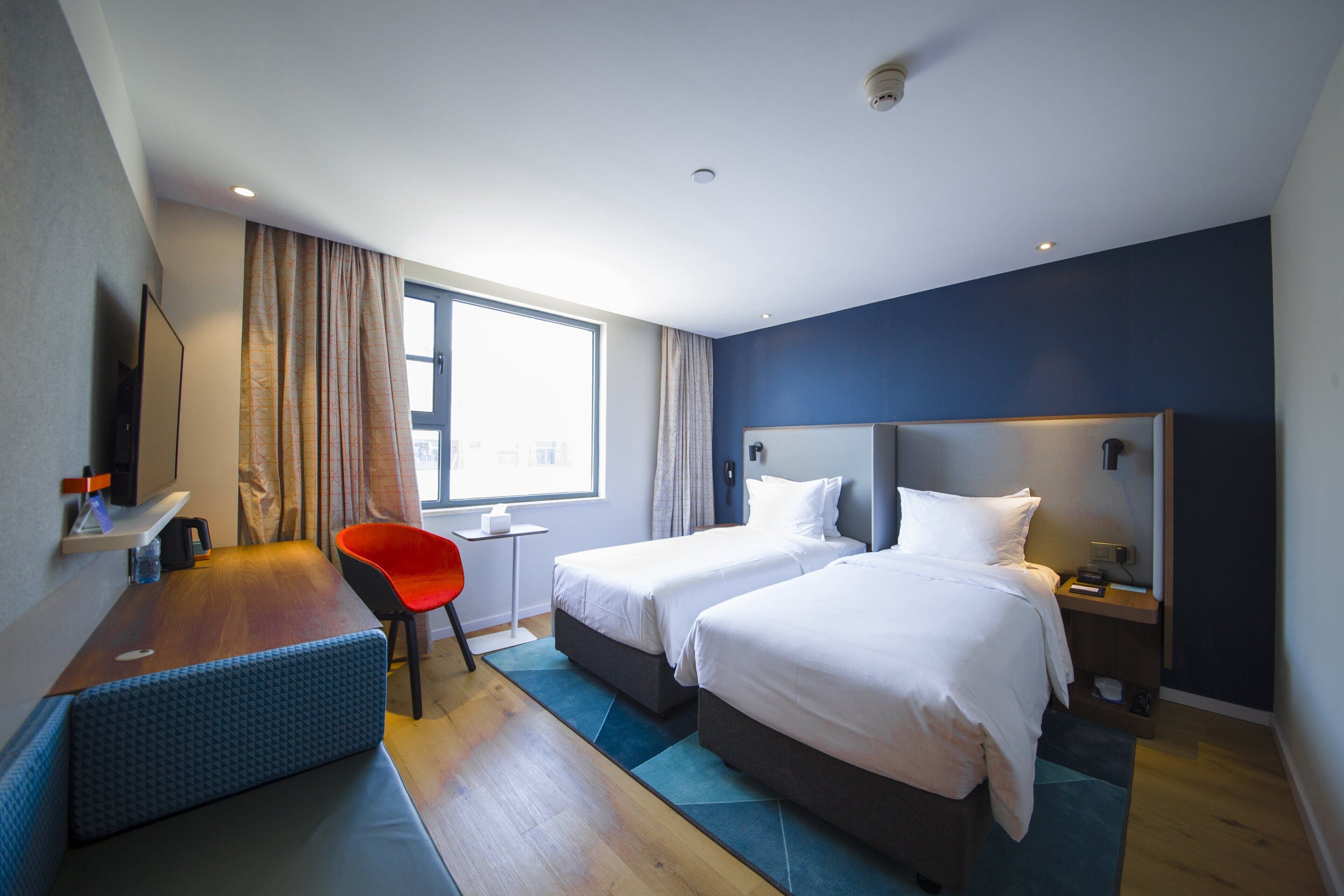 Ein Hotelzimmer mit zwei Betten, einem Schreibtisch, einem Stuhl, einem Fernseher und einem Fenster mit Vorhängen.
