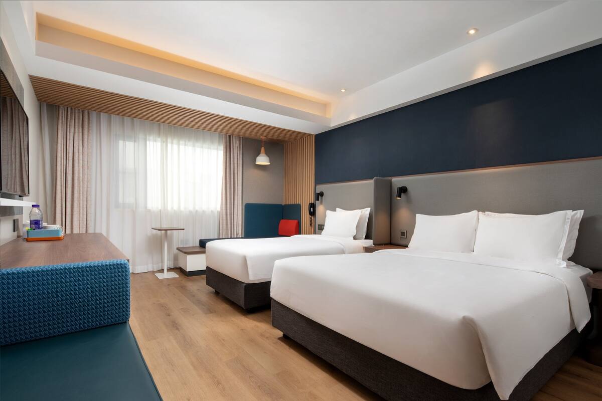 A hotel room with two beds, a wooden floor, a blue bench, and a small table with a bottle and a box.