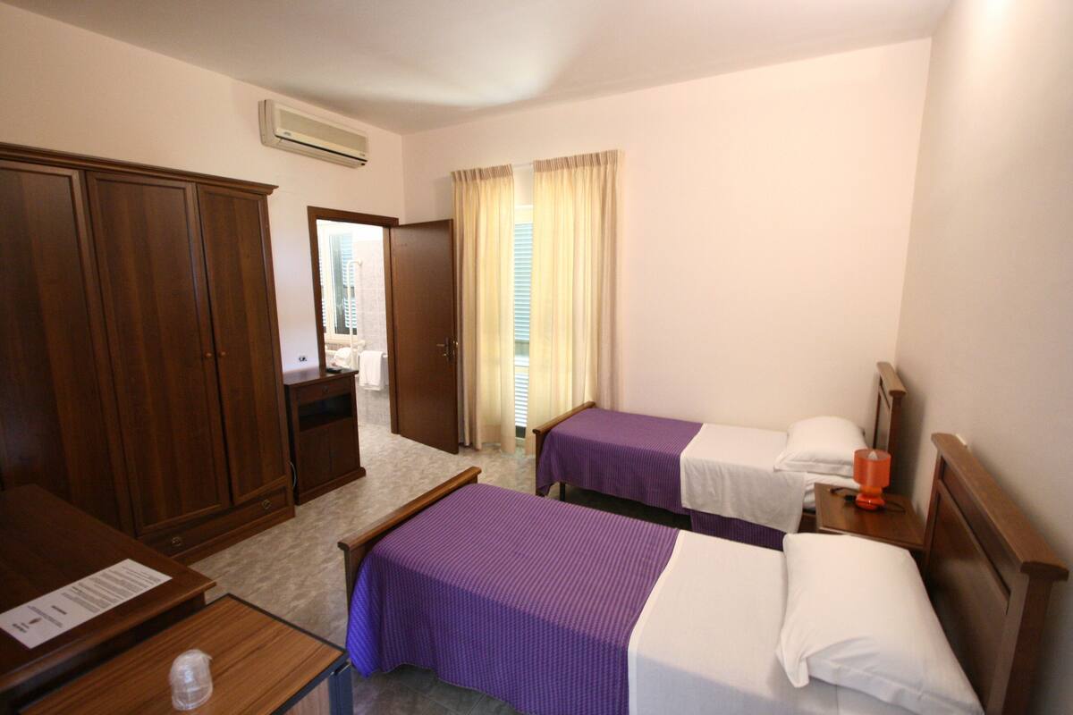 Habitación de hotel con dos camas individuales, un armario de madera, un escritorio con silla y una ventana con cortinas.