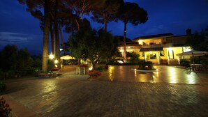 A well-lit outdoor area with a paved walkway, trees, and a building with a balcony. - La Villa Del Colle (Monte San Giovanni Campano)