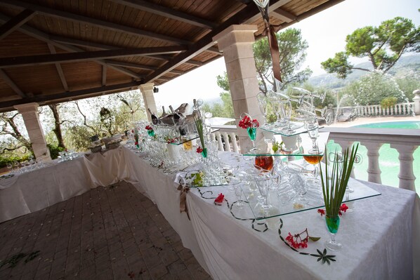 A covered outdoor dining area with a long table set for a meal, surrounded by trees and a pool. - La Villa Del Colle (Monte San Giovanni Campano)