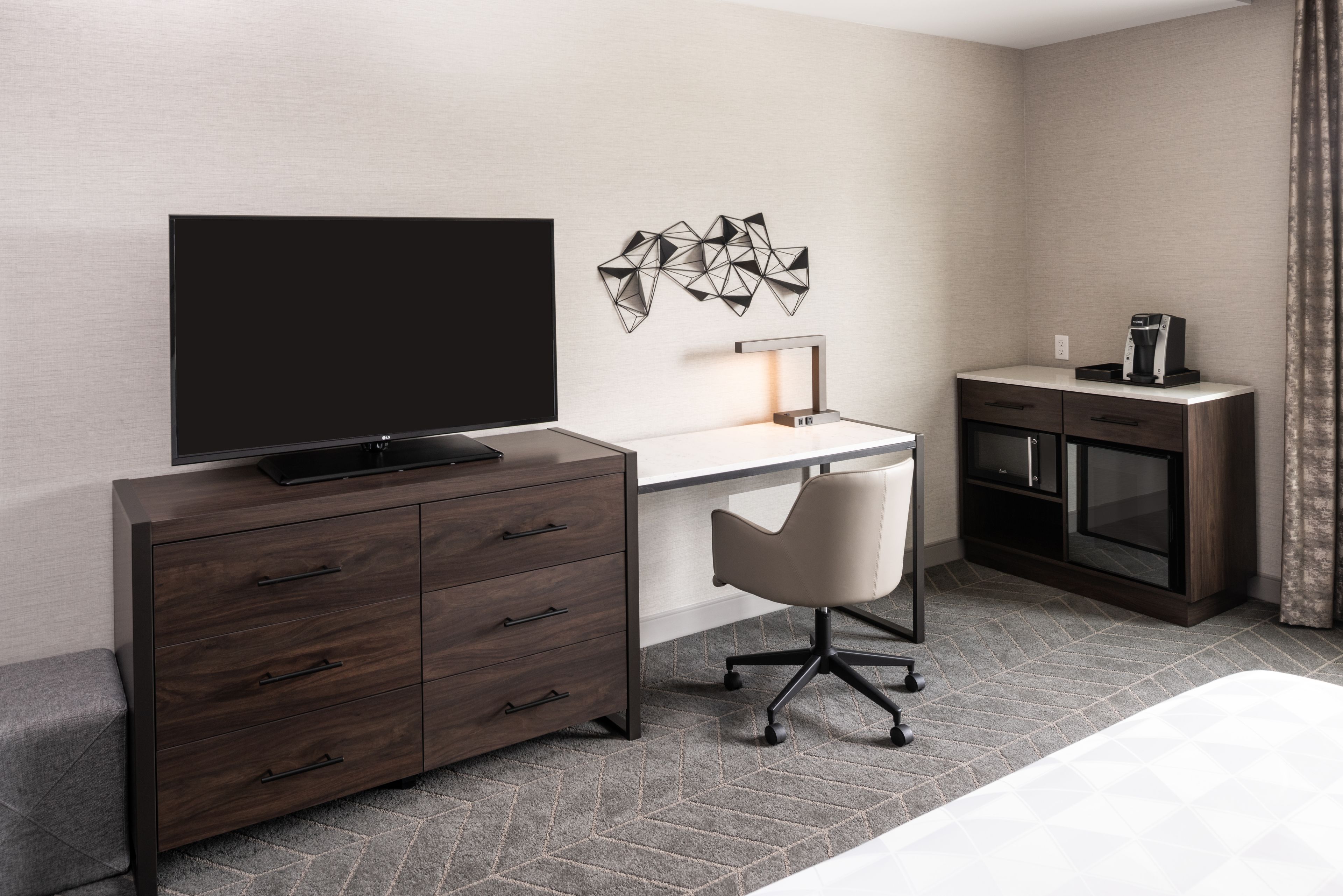 A modern hotel room with a large flat-screen TV, a desk with a chair, a coffee machine, and a minibar.