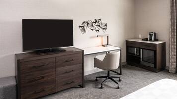 Quarto de hotel moderno com televisão de tela plana grande, uma mesa com cadeira, máquina de café e minibar.