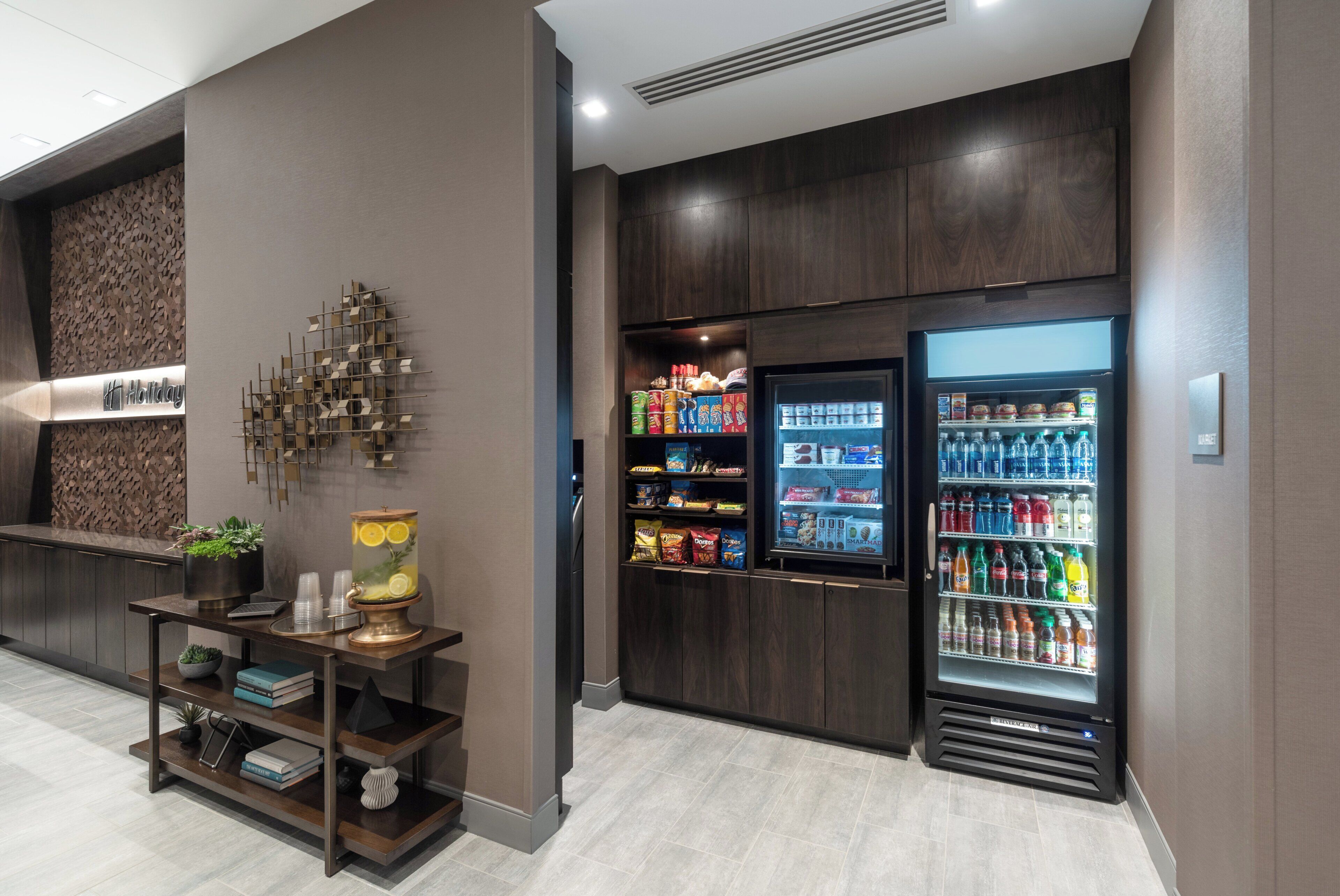 A modern hotel lobby with a reception desk, a decorative wall piece, a beverage dispenser, and a small seating area with books and a plant.