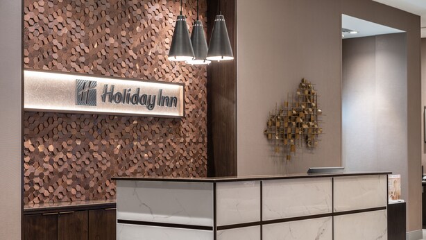 A reception desk with a sign reading "Holiday Inn" and a decorative wall with a geometric pattern. - Holiday Inn Boston Logan Airport - Chelsea by IHG (Chelsea)