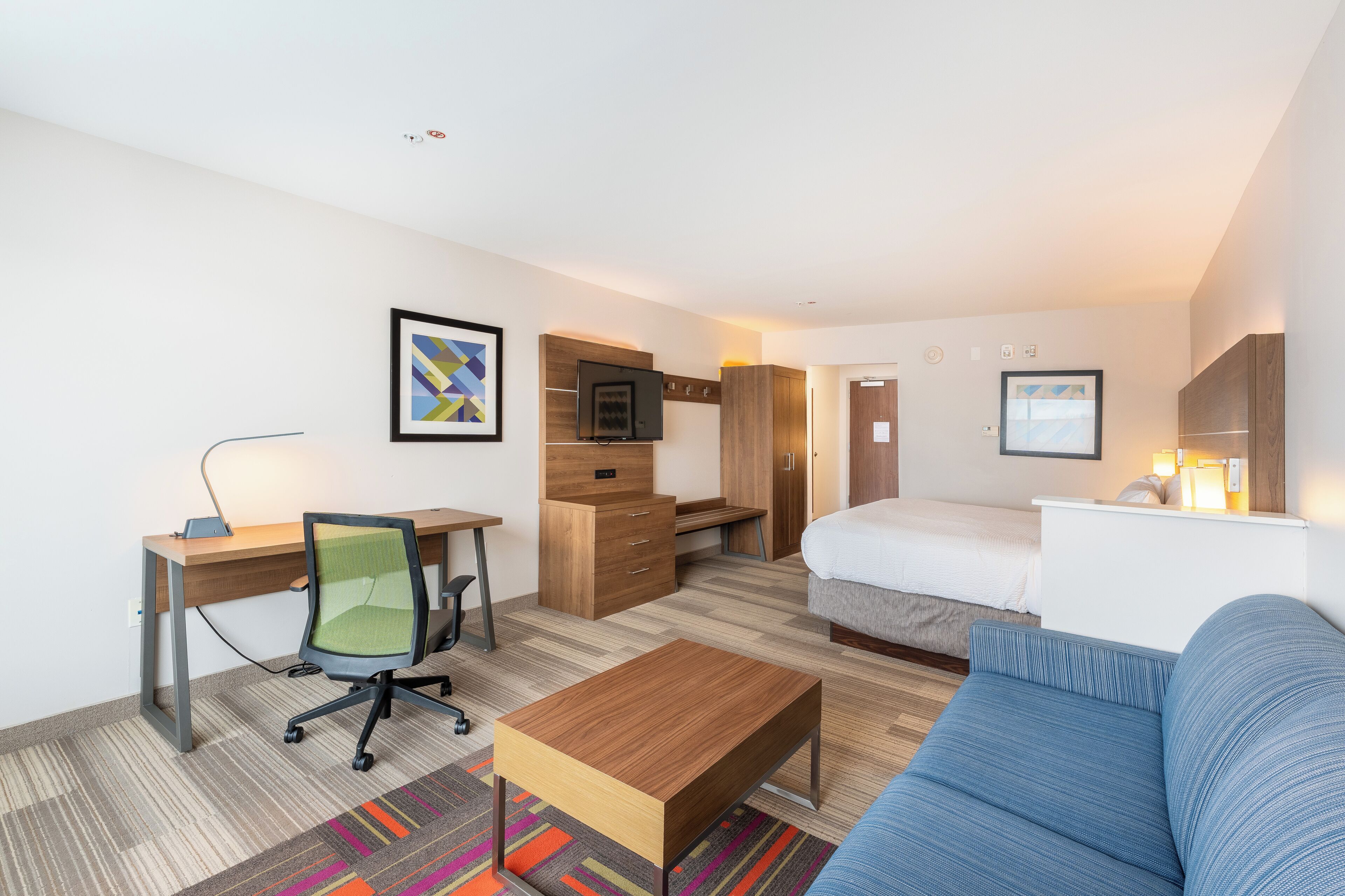 A hotel room with a bed, a desk with a chair, a TV, a sofa, and a colorful rug.
