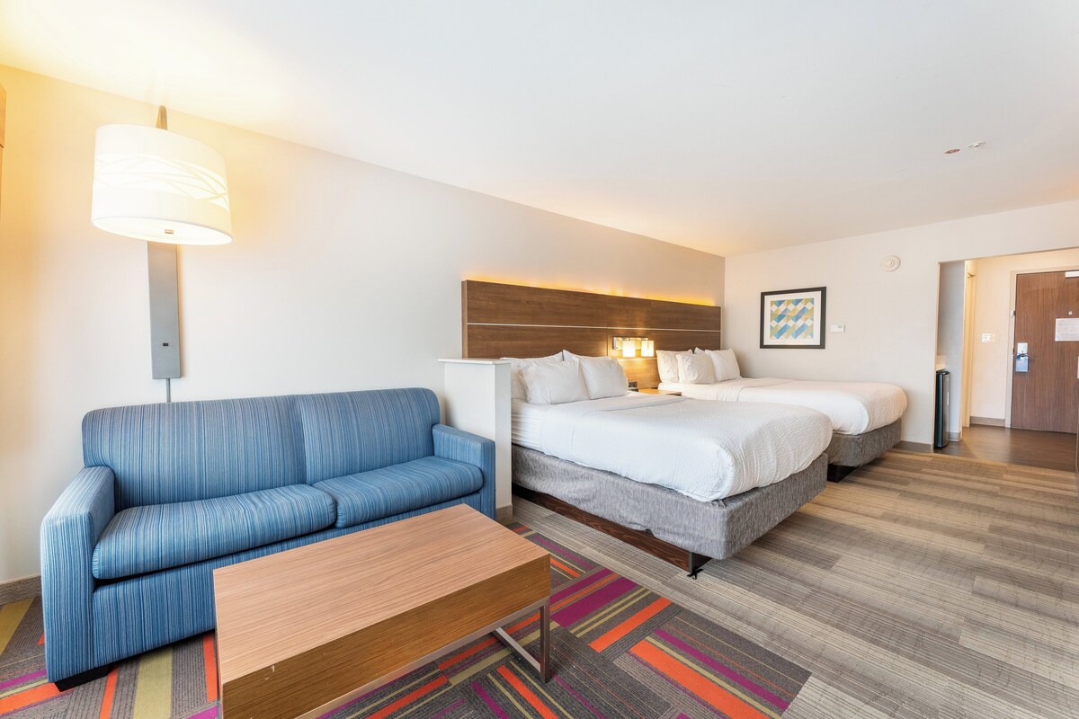 A hotel room with a blue sofa, a wooden coffee table, a bed with two pillows, and a colorful striped rug.