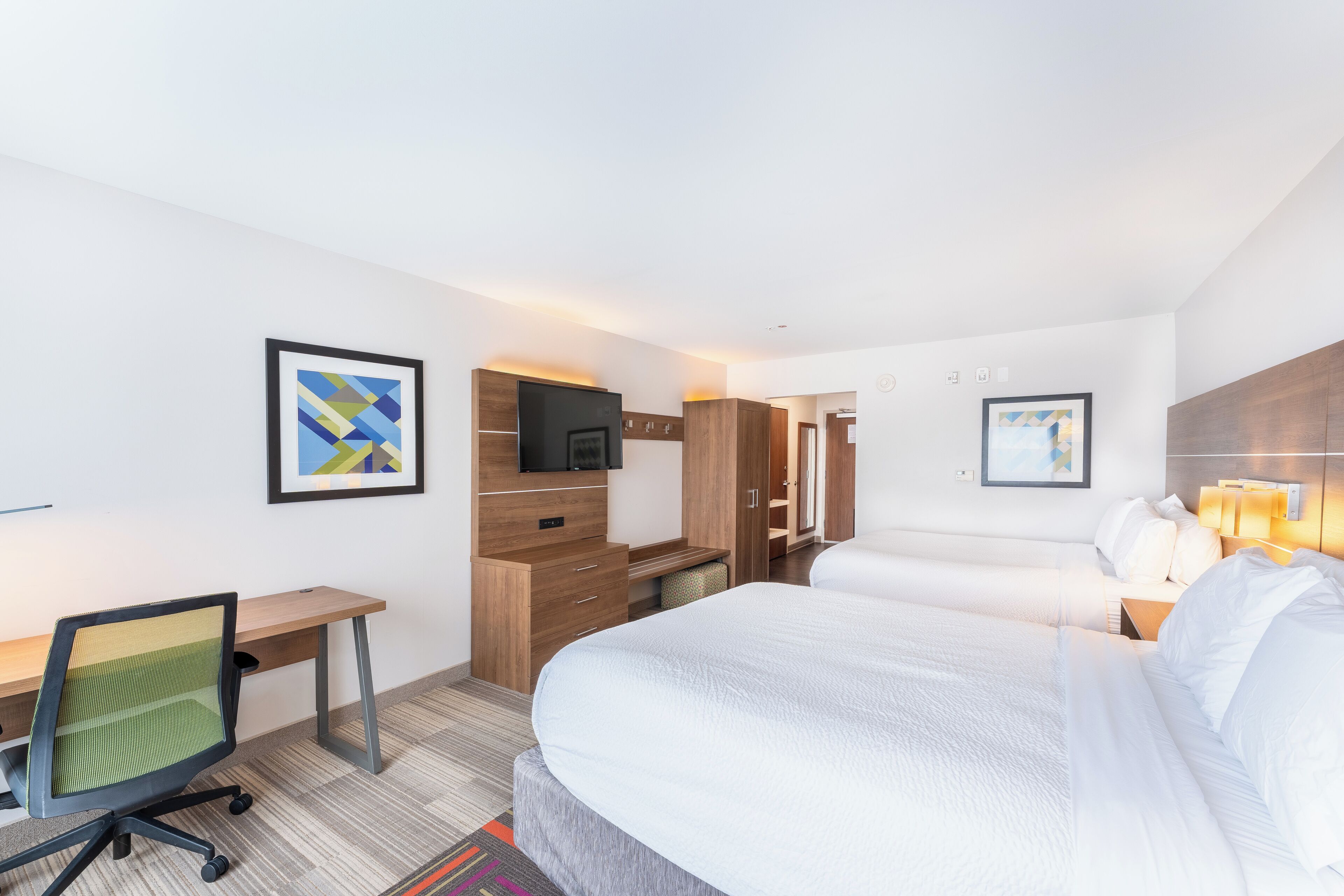 A hotel room with a bed, desk, chair, TV, and artwork.