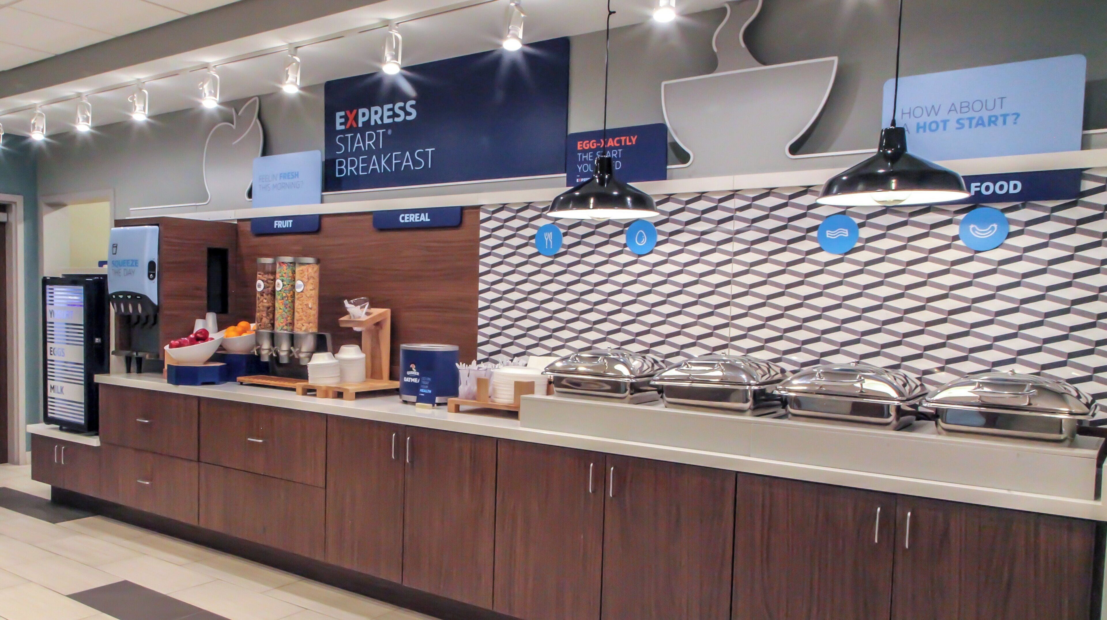 Espaço de buffet de café da manhã com estações de comida identificadas, dispensador de bebidas e parede com padrão.