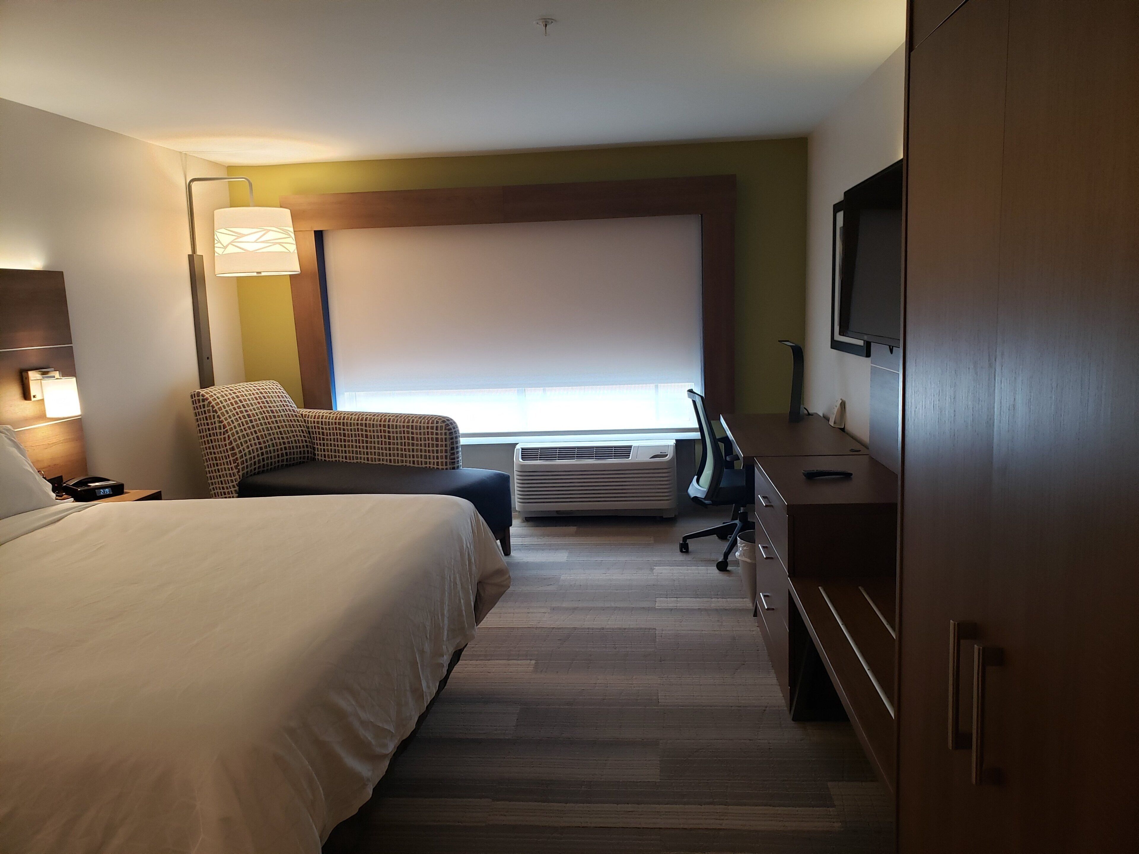 Een hotelkamer met een bed, bureau, stoel, televisie en een raam met luxaflex.