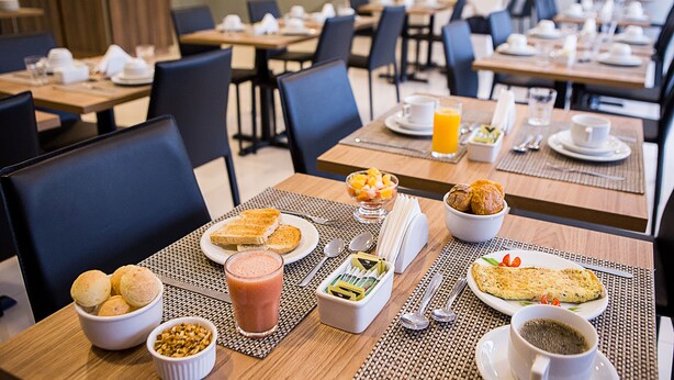 A breakfast setting with tables set for dining, including plates of toast, a bowl of fruit, a smoothie, and a cup of coffee. - Trip Hotel Ubá (Uba)