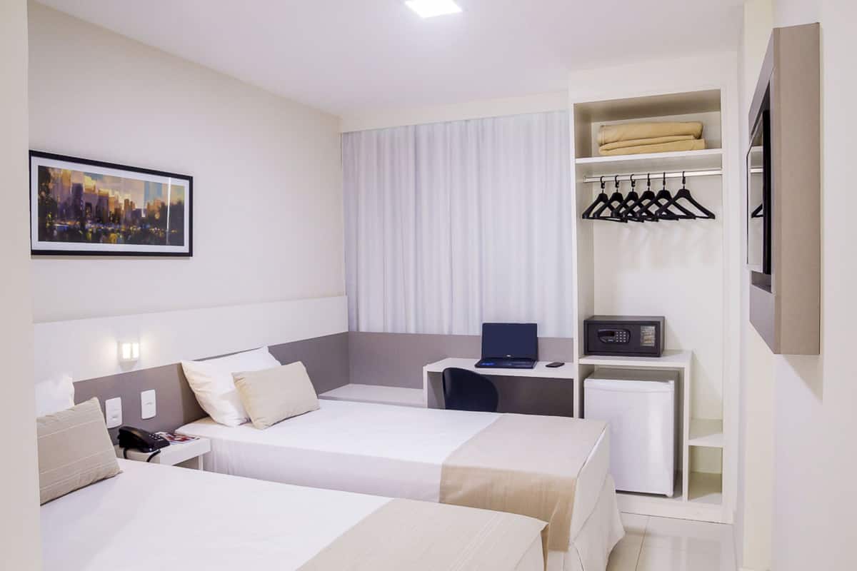 Hotelkamer met twee bedden, een bureau met een laptop, een magnetron en een kleine koelkast. Er staat een kapstok met kleerhangers en er hangt een lijst aan de muur.