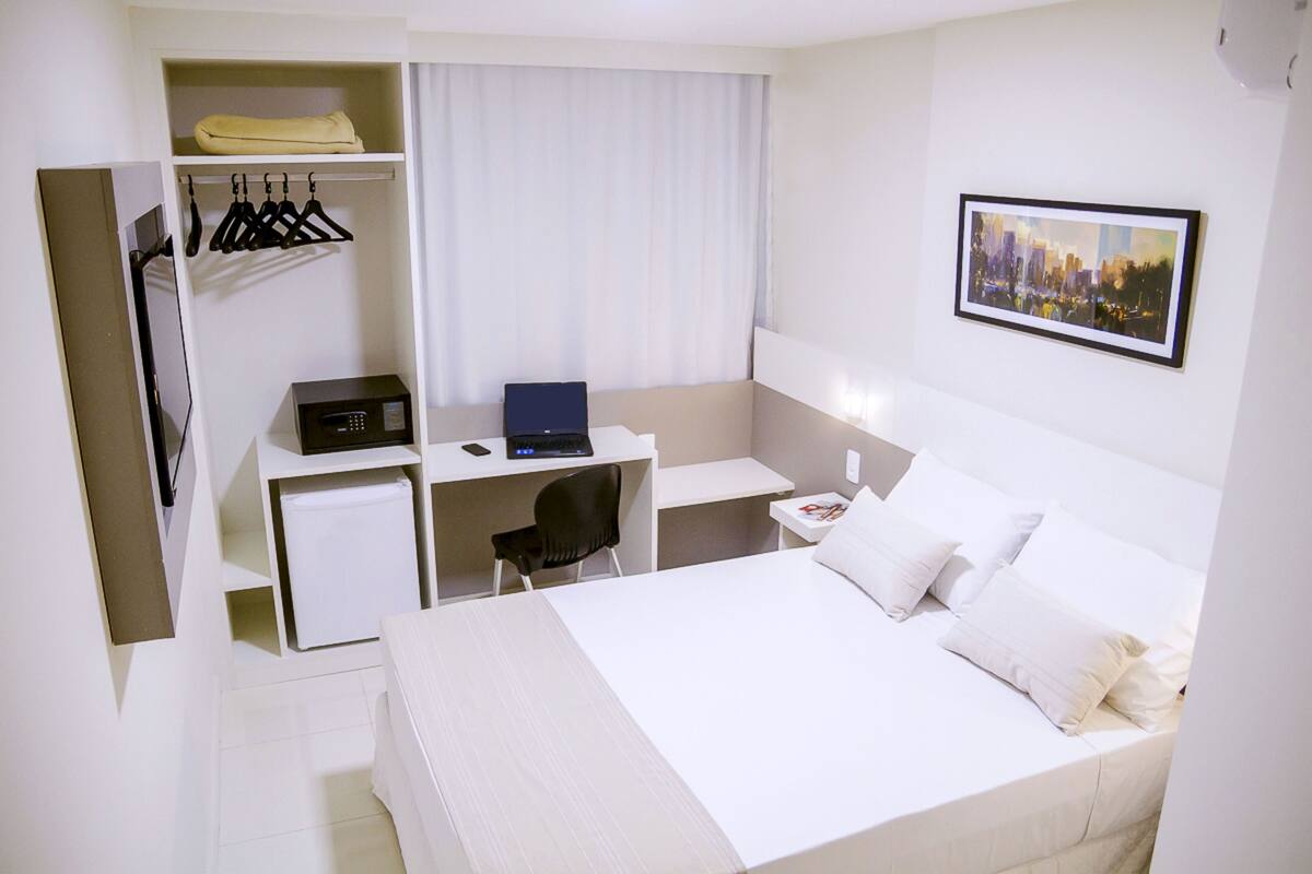 Een hotelkamer met een bed, een bureau met een computer, een stoel, een kleine koelkast, een kledingkast en een stadsgezichtschilderij aan de muur.