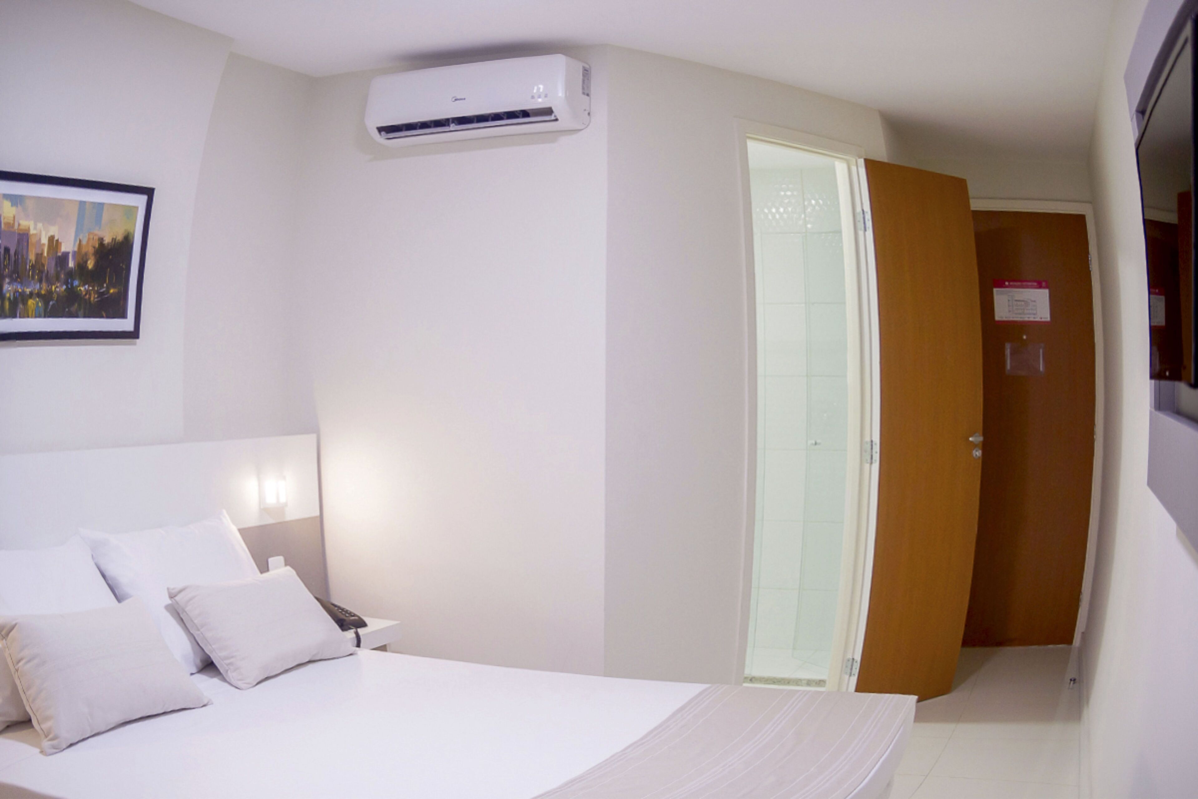 A hotel room with a bed, white walls, an air conditioning unit, a painting, and a wooden door.