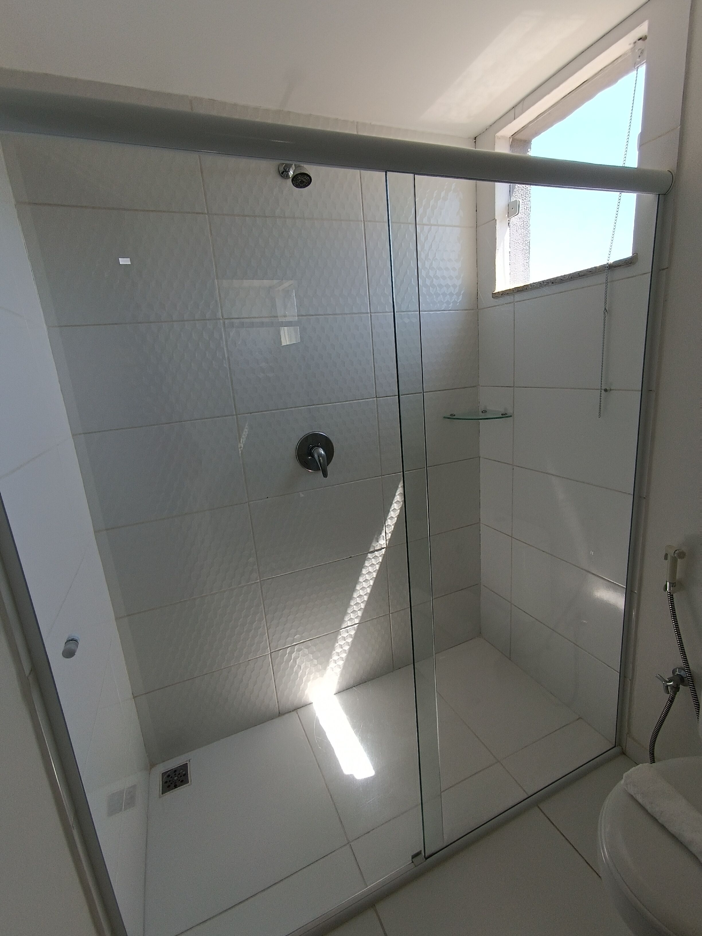 Zona de ducha con paredes de cristal, suelo de baldosas y una ventana arriba.