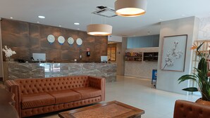 A lobby with a brown leather sofa, a wooden coffee table, and a reception desk with multiple clocks. - Trip Hotel Ubá (Uba)