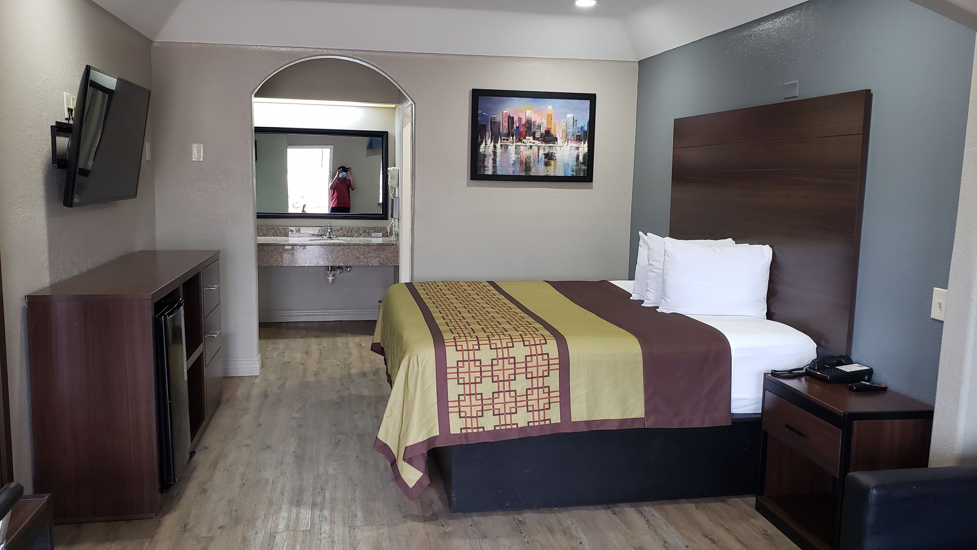 A hotel room with a bed, a desk, a mirror, a painting, and a view of the cityscape.