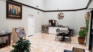 A compact kitchen with a coffee machine, a dining table with chairs, a potted plant, and a travel-themed bulletin board. - Palace Inn Hwy 6 North & Clay Rd (Houston)