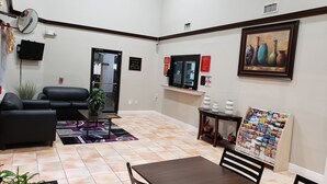 A hotel lobby with a black leather sofa, a small table with a plant, a tiled floor, a television, a framed painting, and a magazine rack. - Palace Inn Hwy 6 North & Clay Rd (Houston)