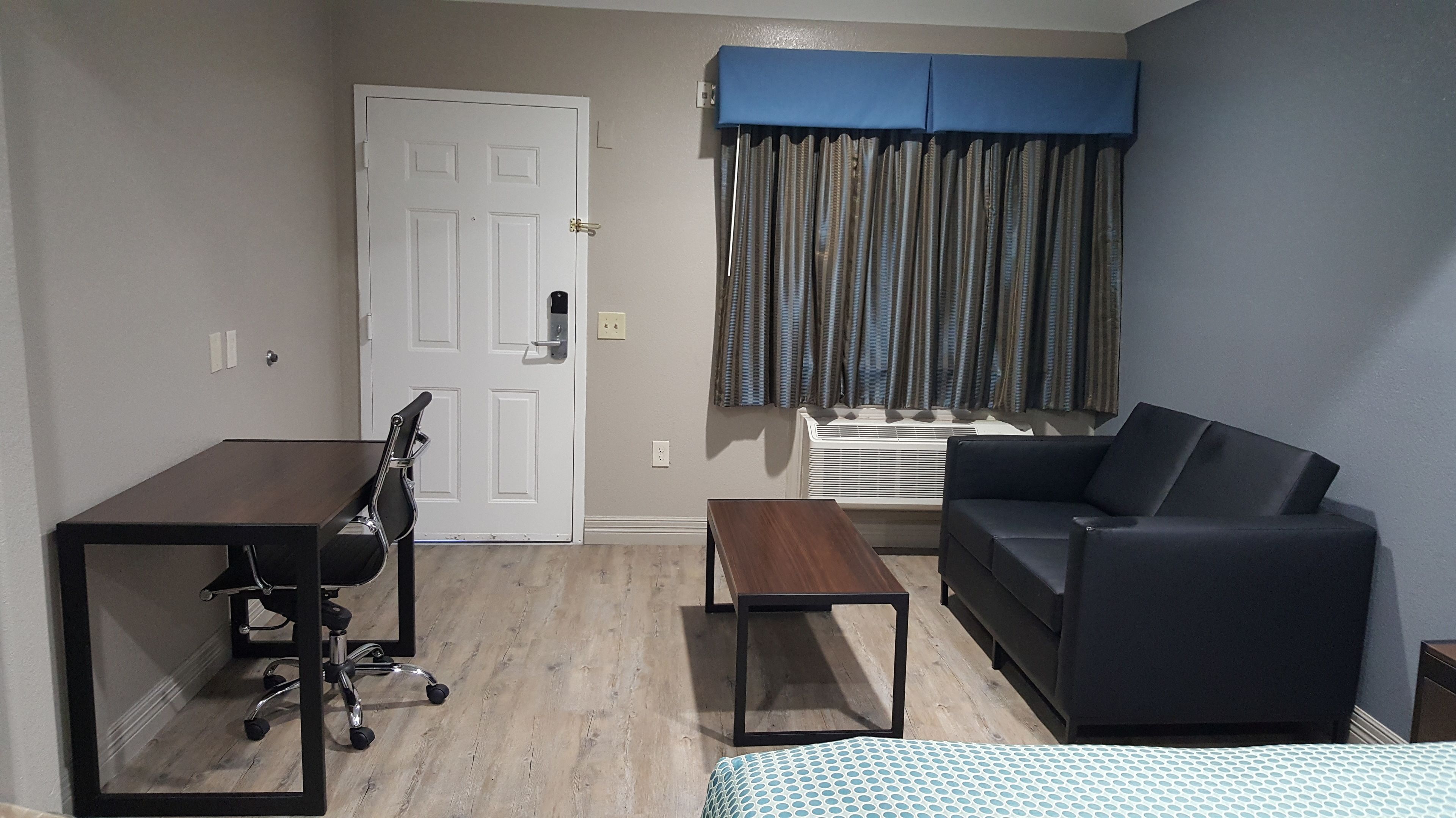 A compact room with a door, a window with curtains, a black sofa, a small table, and a chair with a laptop.