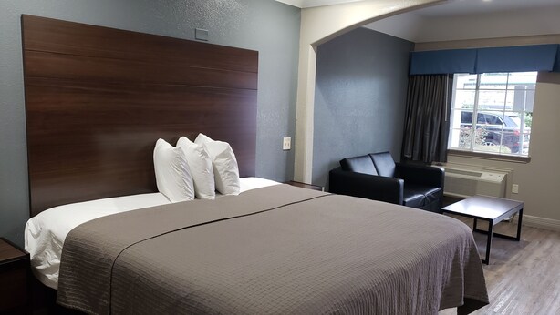 A hotel room with a large bed, a wooden headboard, a black leather chair, a small table, and a window with curtains. - Palace Inn Hwy 6 North & Clay Rd (Houston)