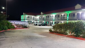 A multi-story hotel with a red roof and green exterior lighting. There are several parked vehicles in front, and a well-maintained garden area with a red curb and "No Parking" sign. - Palace Inn Hwy 6 North & Clay Rd (Houston)