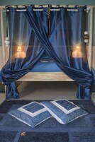 Una cama con una colcha azul y almohadas a juego, una cortina azul y una alfombra pequeña con un diseño geométrico.