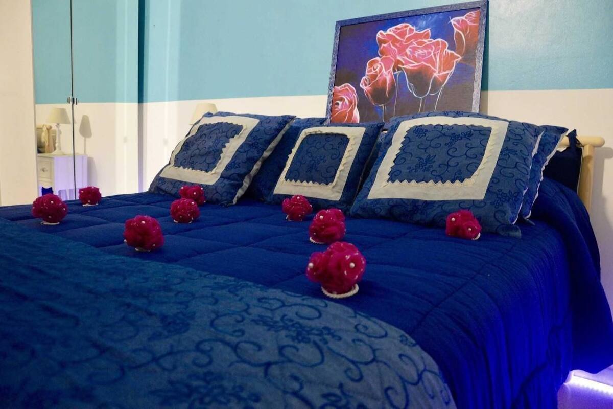 Una cama con colcha azul y almohadas decorativas, un cuadro con rosas en la pared y un espejo a la izquierda.