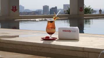 A glass of iced tea with a straw and a decorative fruit on a red plate, a white towel with "Intercity Hotel" text, and a cityscape view in the background.