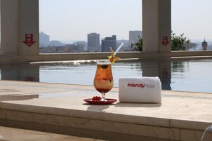 A glass of iced tea with a straw and a decorative fruit on a red plate, a white towel with "Intercity Hotel" text, and a cityscape view in the background.