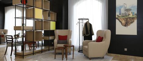 A modern living room with a high ceiling, a large window with sheer curtains, a beige armchair with red cushions, a wooden bookshelf, and a coat rack with clothes.