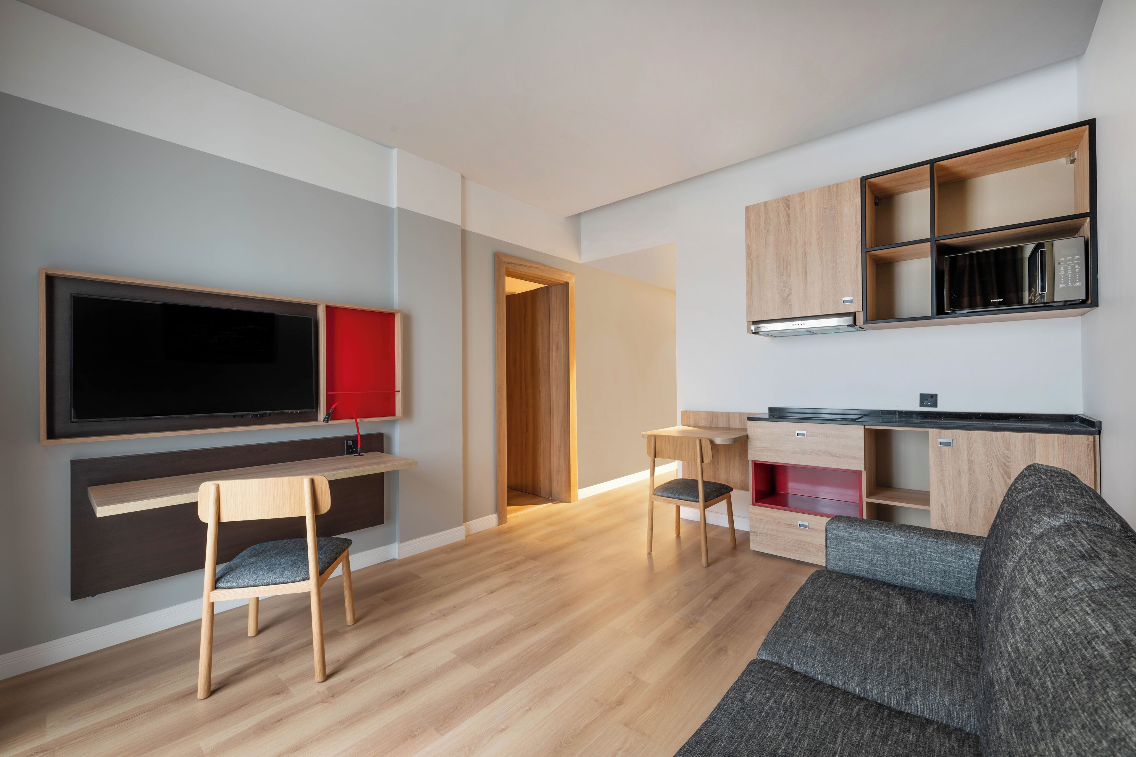A modern living room with a flat-screen TV, wooden furniture, a grey sofa, and a kitchen area with wooden cabinets and a microwave.